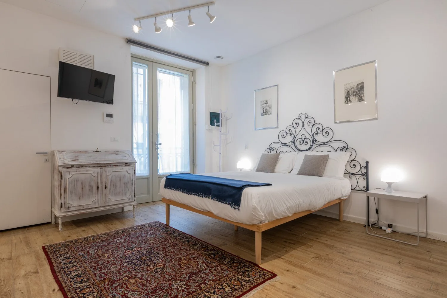 Un dormitorio luminoso con una cama, una alfombra estampada y ventanas altas.