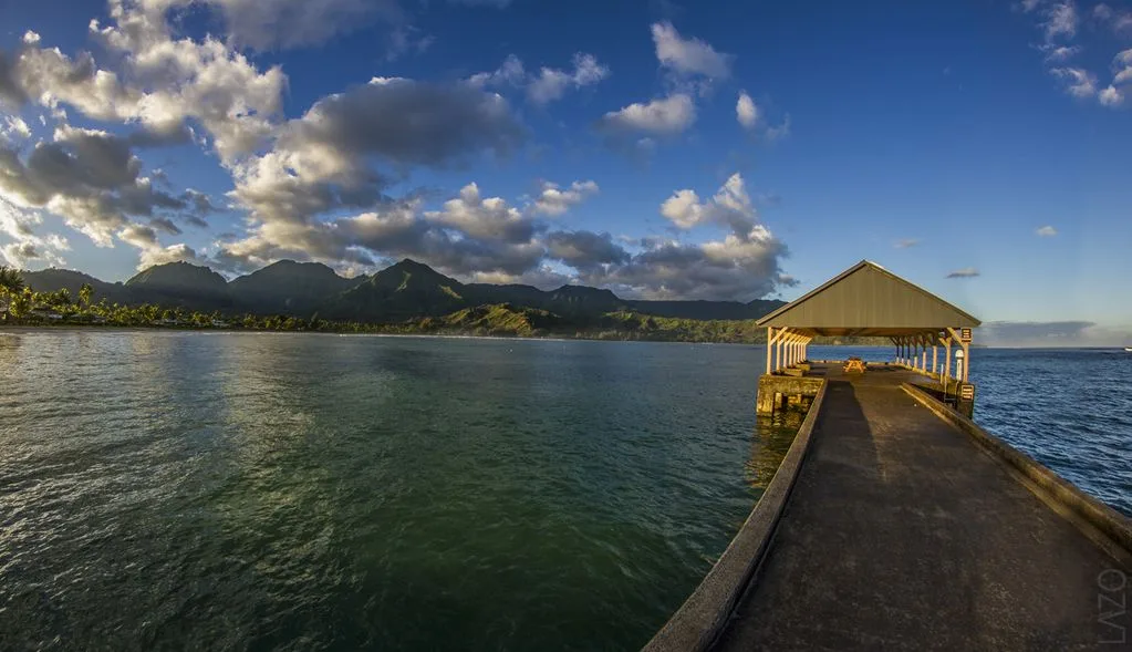 A covered pier extends over the water toward a mountain-lined shore beneath a cloudy sky.