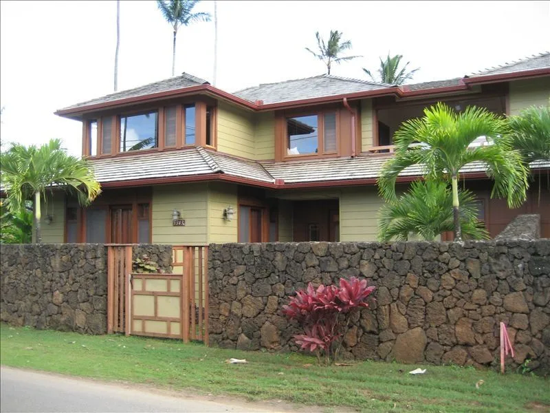 A two-story house with a tiled roof stands behind a stone wall and tropical plants.