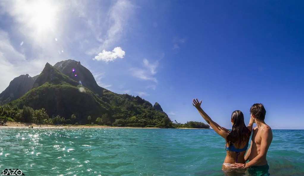 A couple stands in turquoise water beneath a green mountain.