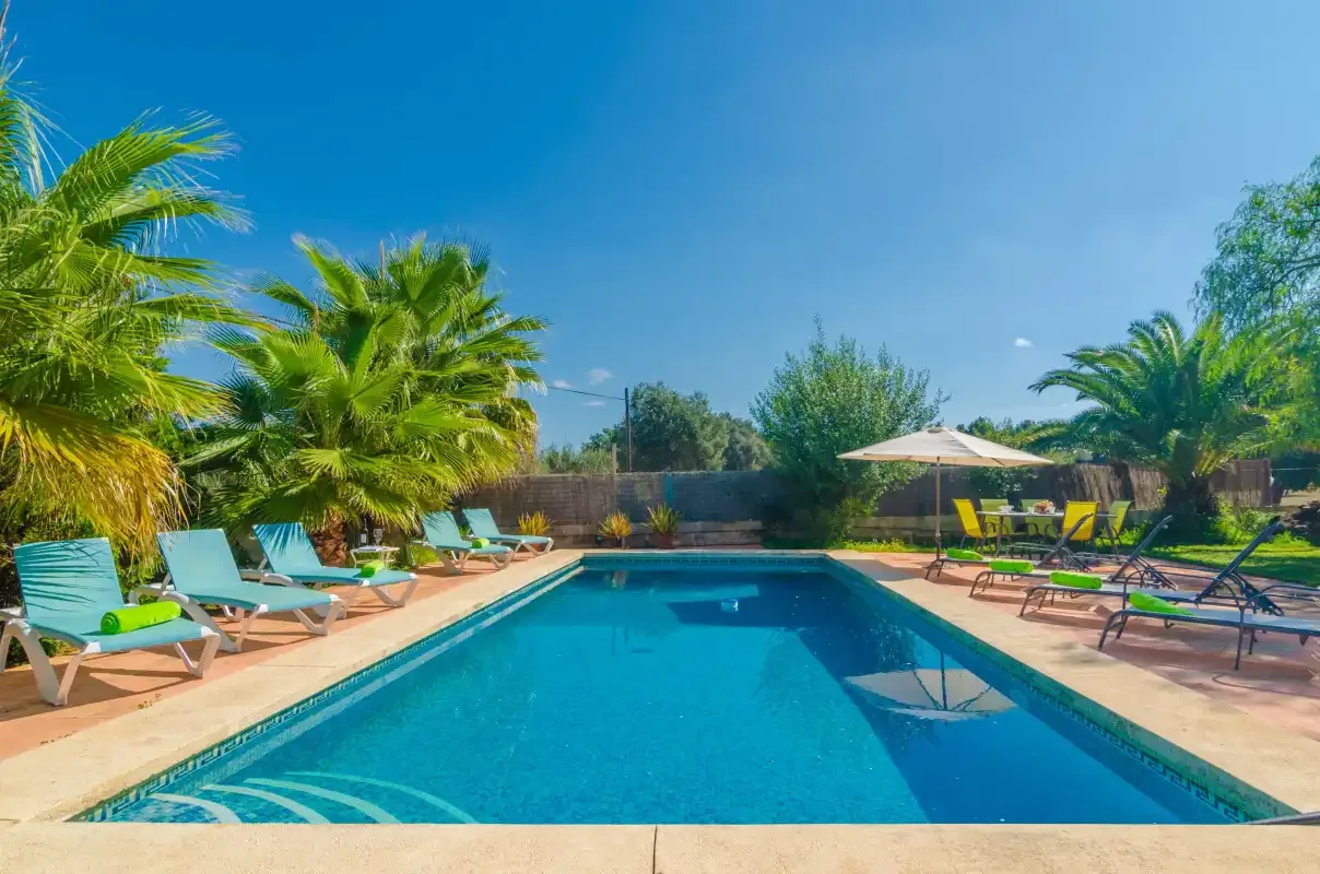 An outdoor swimming pool is surrounded by lounge seating, palm trees, and an umbrella.