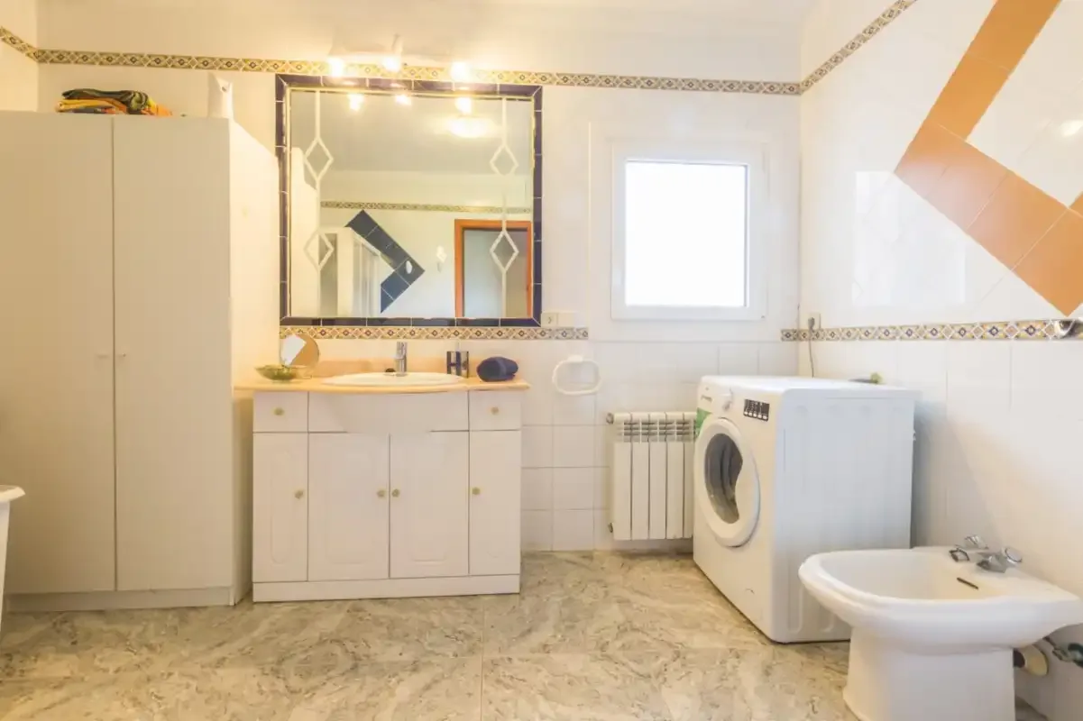 A bright bathroom with a vanity, mirror, washing machine, and bidet.