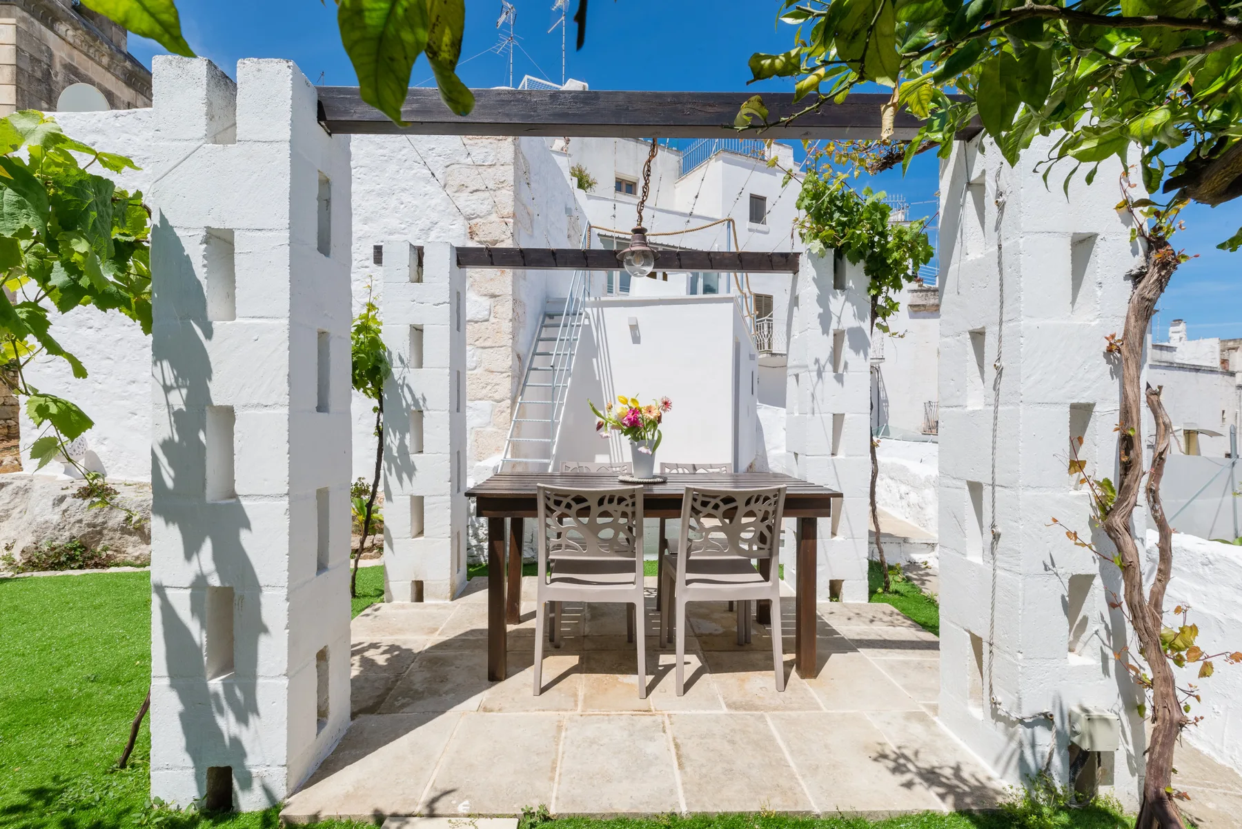 Zona de comedor en un patio de piedra, con mesa y sillas bajo una pérgola de madera.