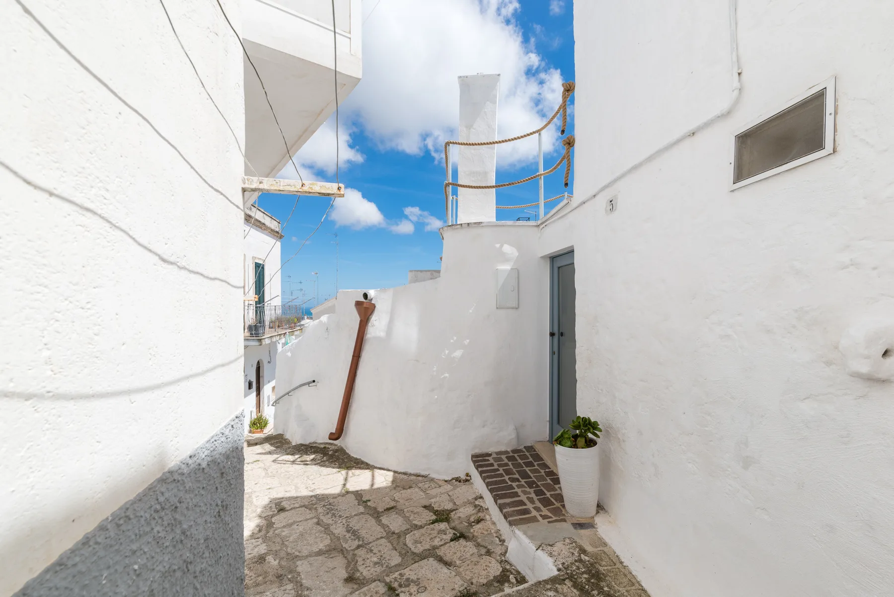 Un pasaje exterior estrecho entre paredes blancas, con pavimento de piedra y cielo azul.
