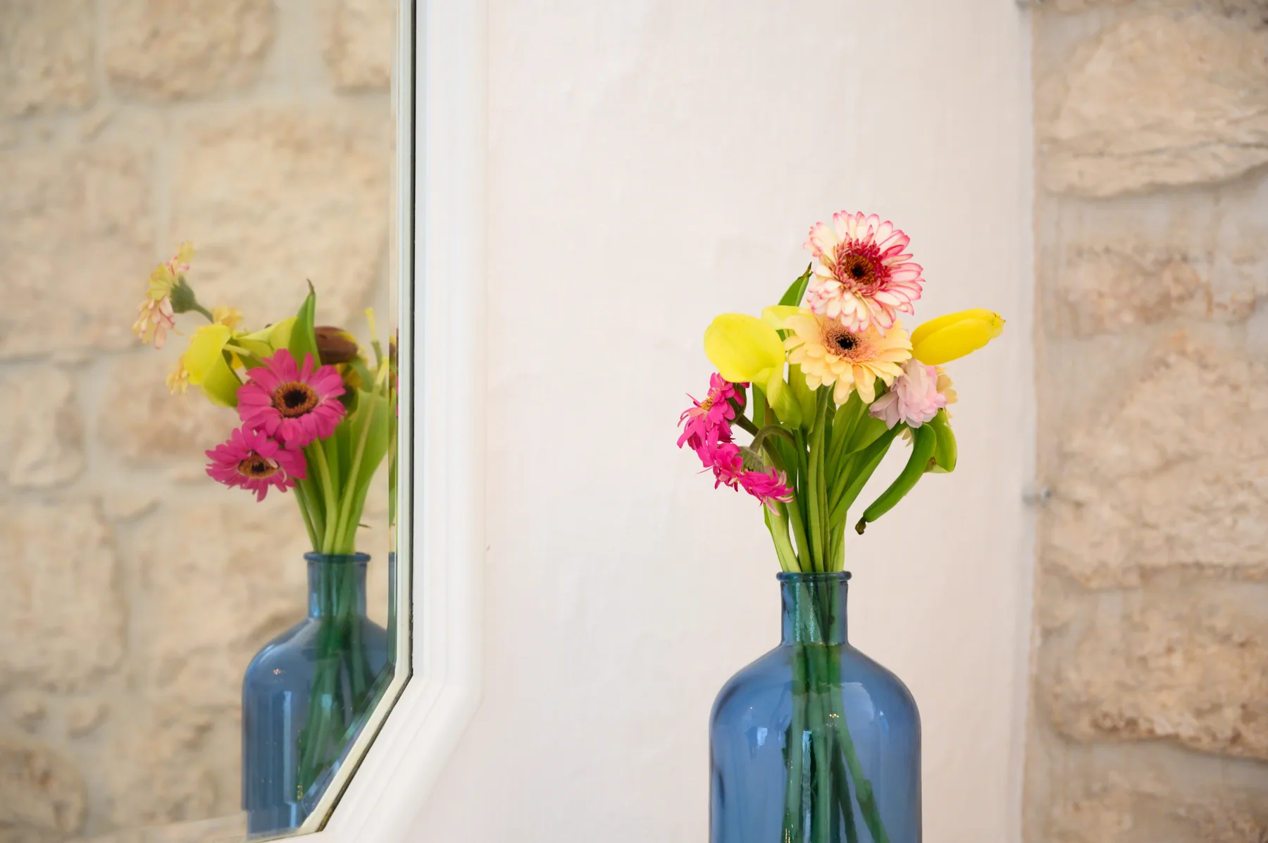 Un jarrón de vidrio azul con flores de colores está junto a un espejo y una pared de piedra.
