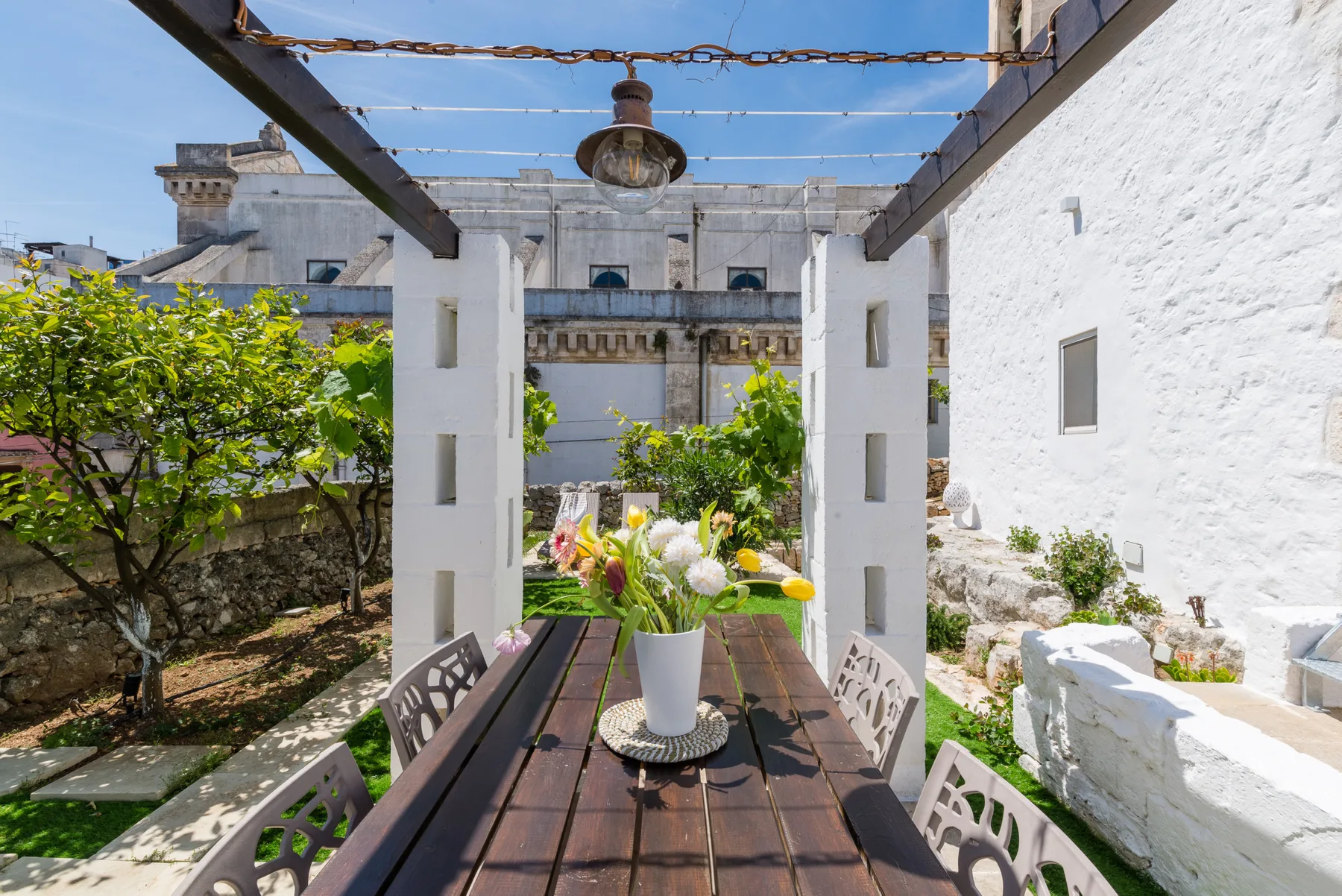 Una zona de comedor al aire libre está bajo una pérgola, con plantas sobre la mesa.