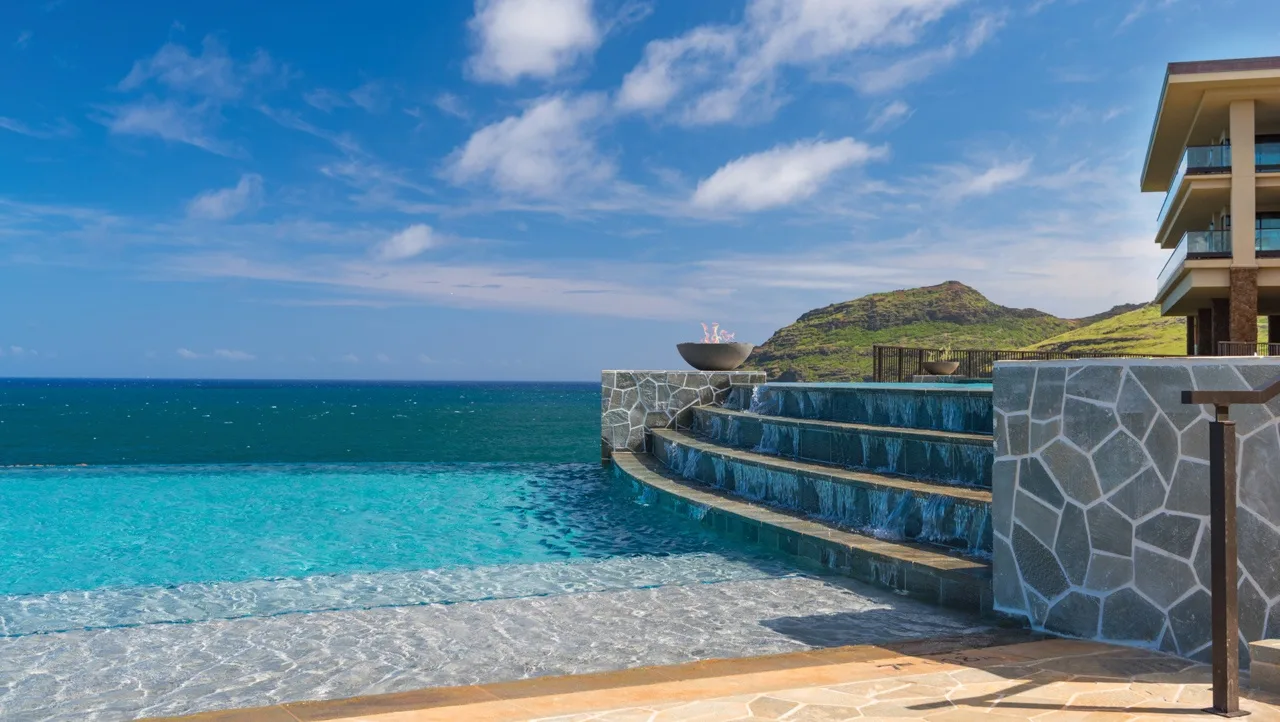 Una piscina infinita da al océano, con un muro de piedra y una colina cercana.