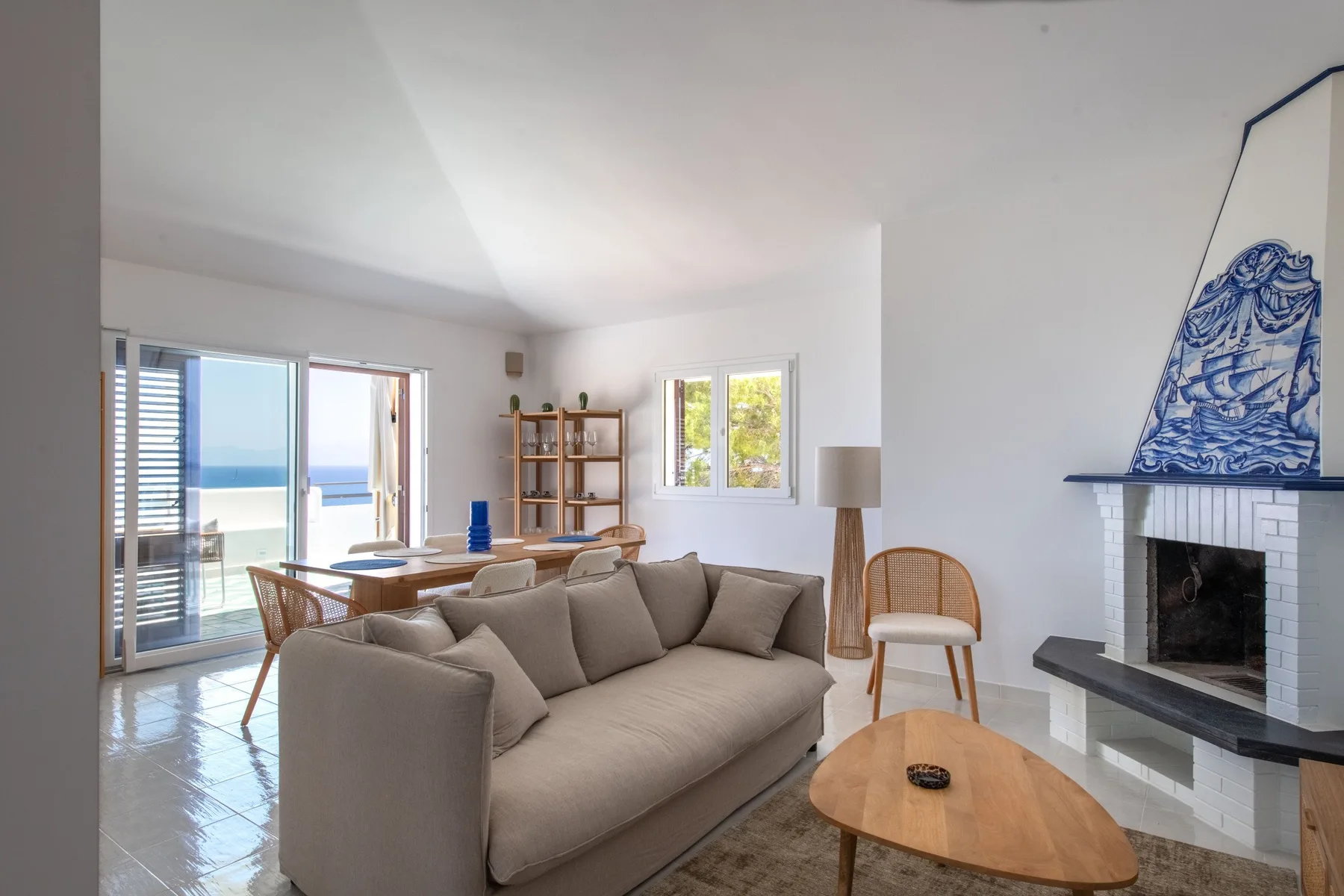 A bright living room with seating, a fireplace, and large windows overlooking the sea.
