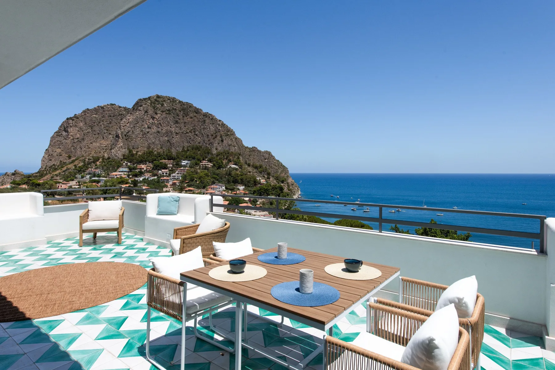 A sunny rooftop terrace has a dining table and seating overlooking the sea and rocky coastline.