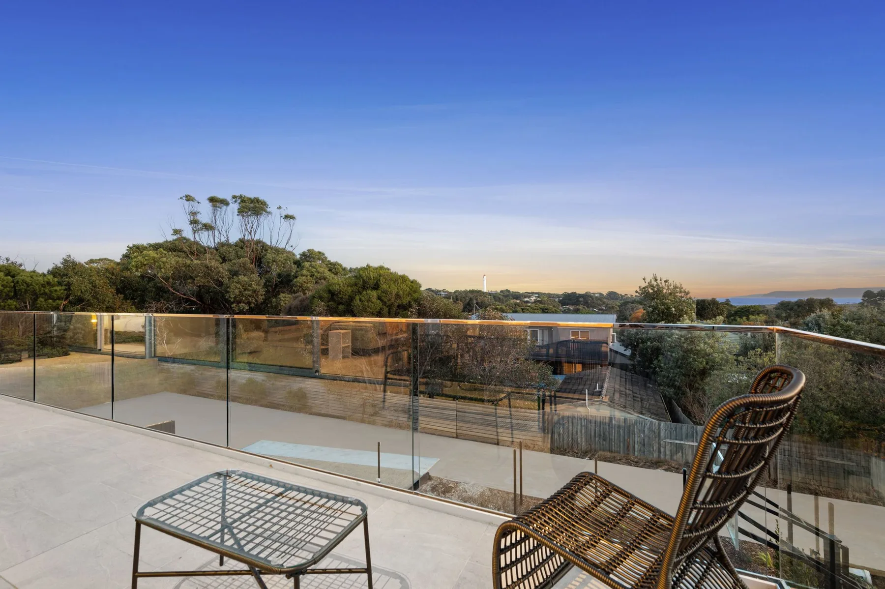 Una terraza soleada da a árboles y agua distante, tras una barandilla de vidrio.