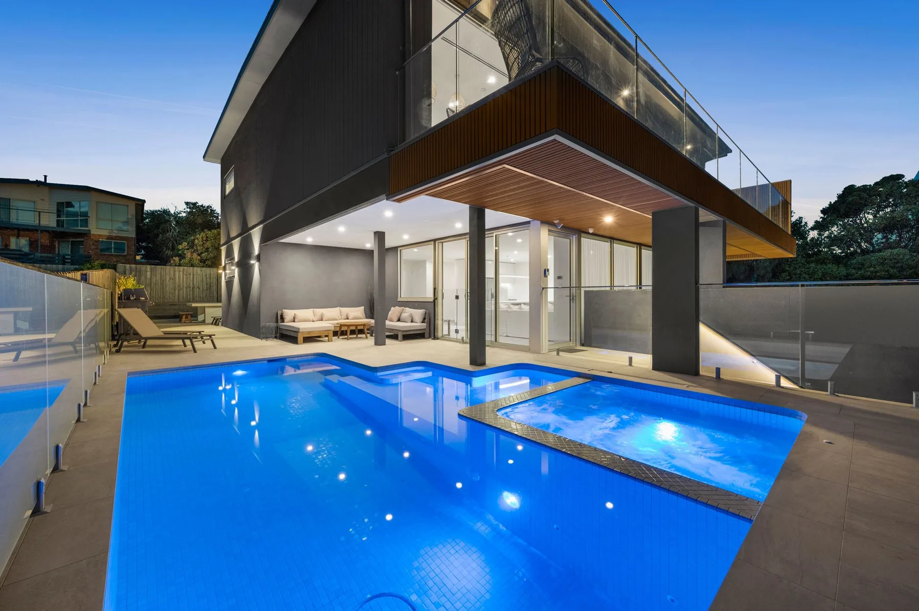 Una piscina exterior iluminada y un spa están junto a una casa moderna.