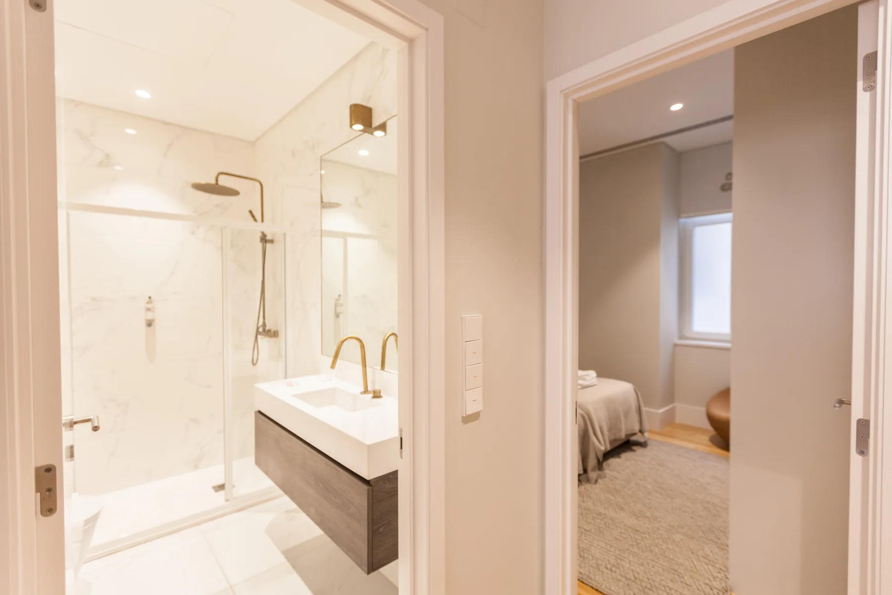 Un baño luminoso con ducha de cristal y lavabo junto a un dormitorio.