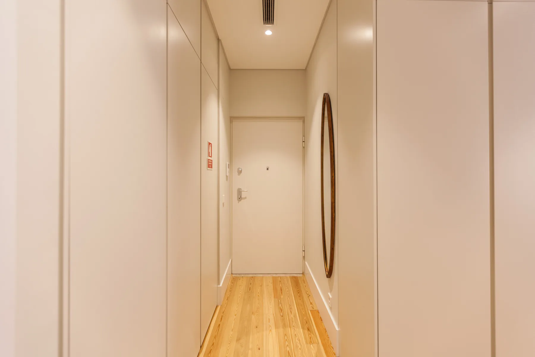 Un pasillo estrecho con suelo de madera clara conduce a una puerta cerrada.