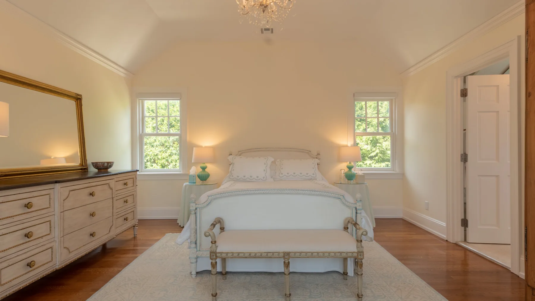A bedroom with a bed, a bench, and a dresser between two windows.