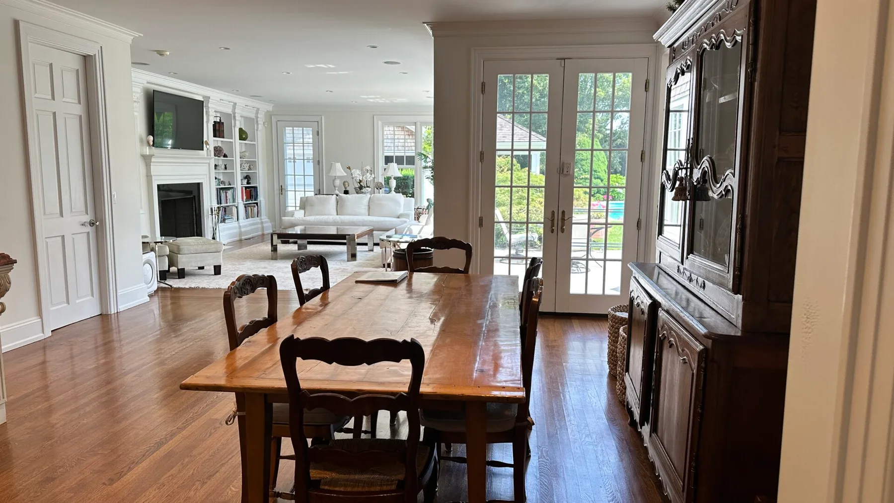 A wooden dining table and chairs sit in a bright room with glass doors.