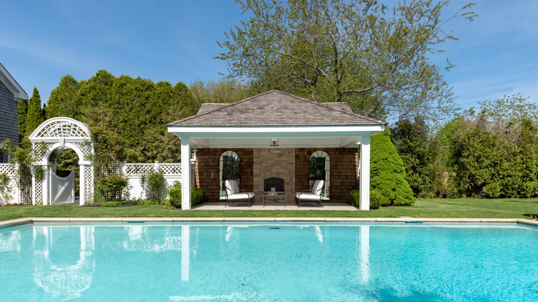 An outdoor swimming pool faces a covered seating area surrounded by greenery.
