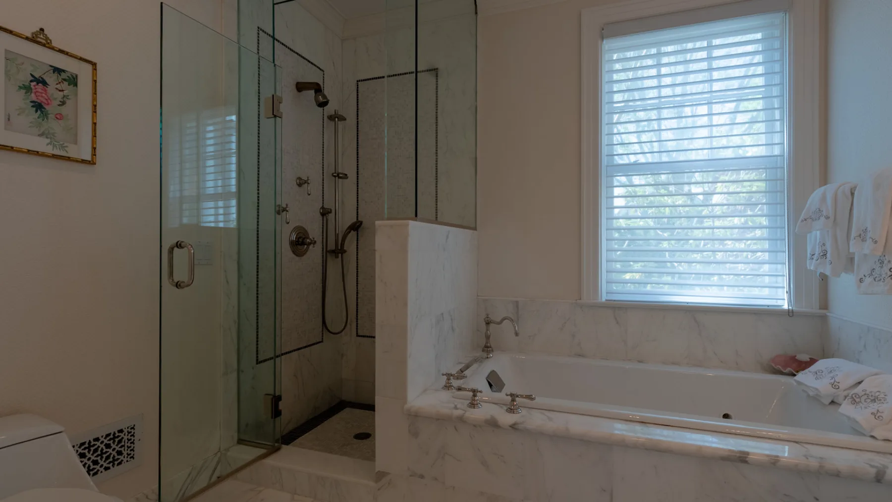 A bathroom with a glass shower, bathtub, and window with blinds.