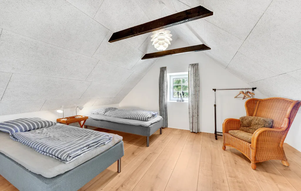 Un dormitorio abuhardillado luminoso con dos camas, una ventana pequeña y asientos de mimbre.