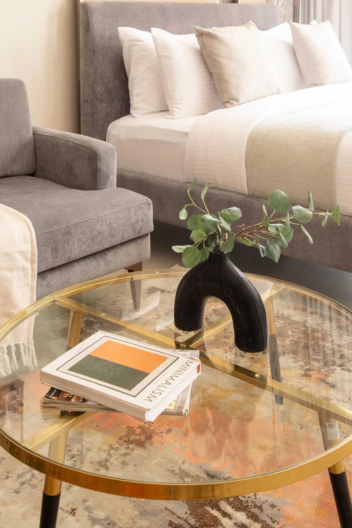 A bedroom with a neatly made bed, gray seating, and a glass coffee table.