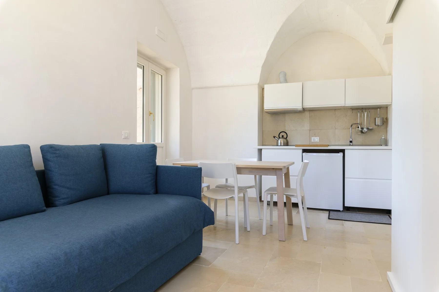 A bright living area with blue seating and a compact kitchen behind a dining table.