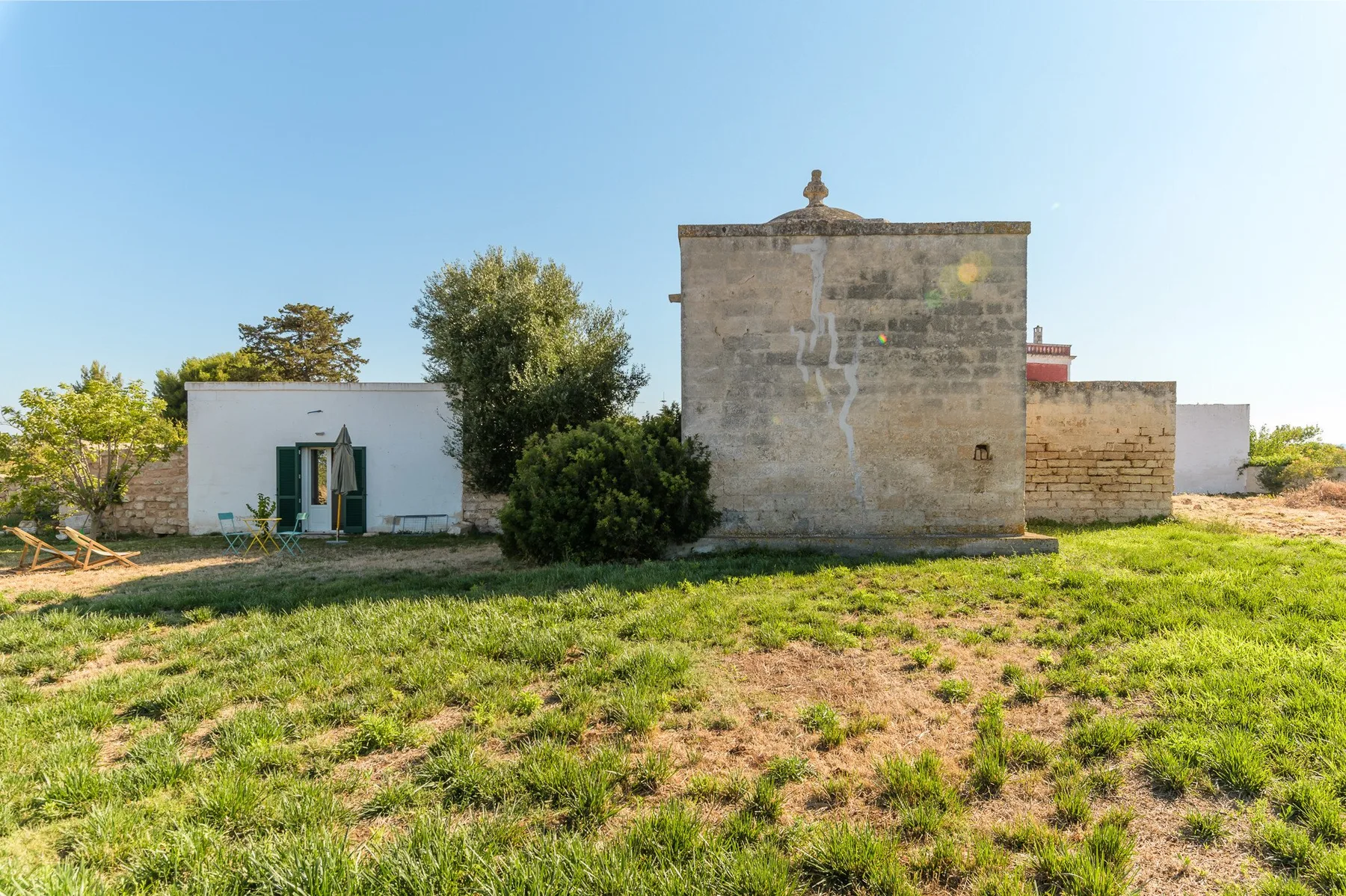 A low stone building and small white structures stand amid grassy ground.
