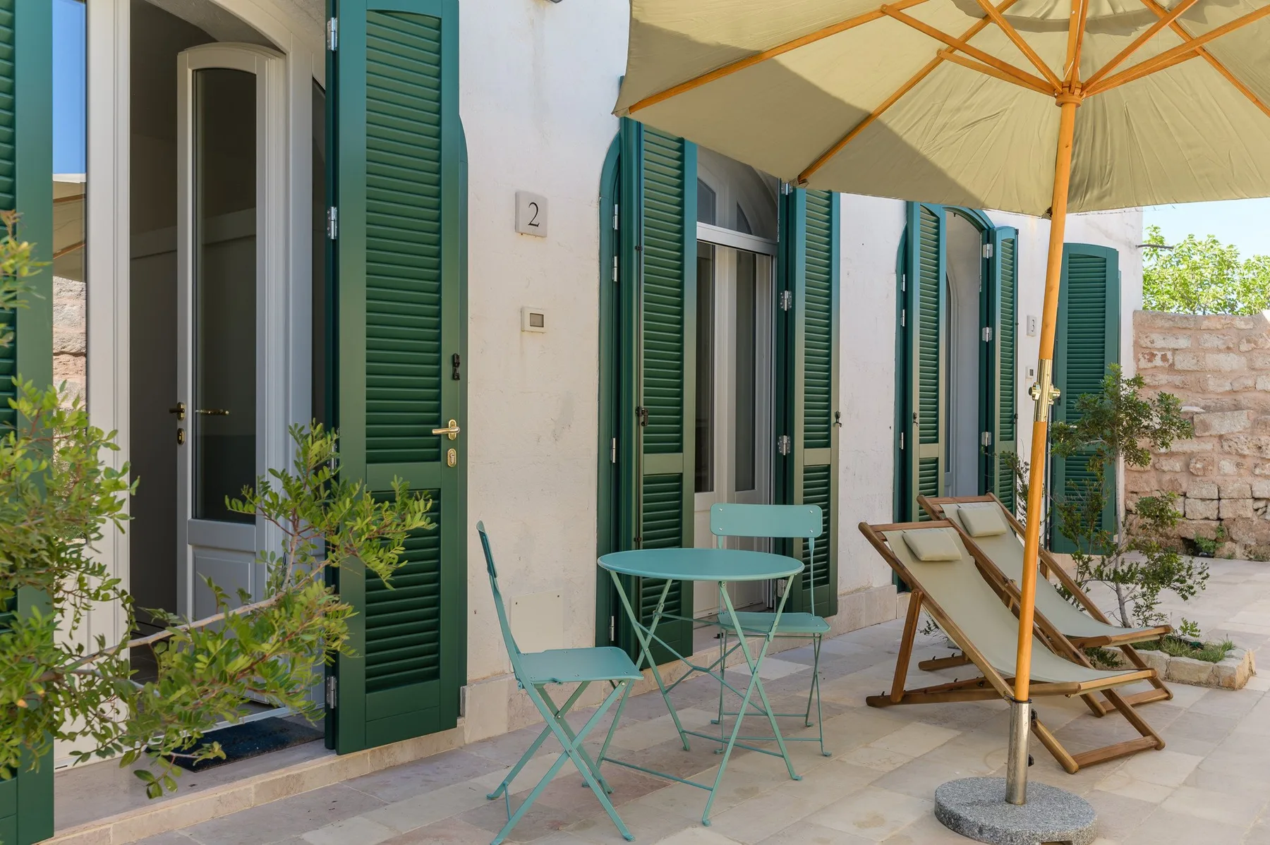 A sunlit patio with green shutters, outdoor seating, and a large umbrella.
