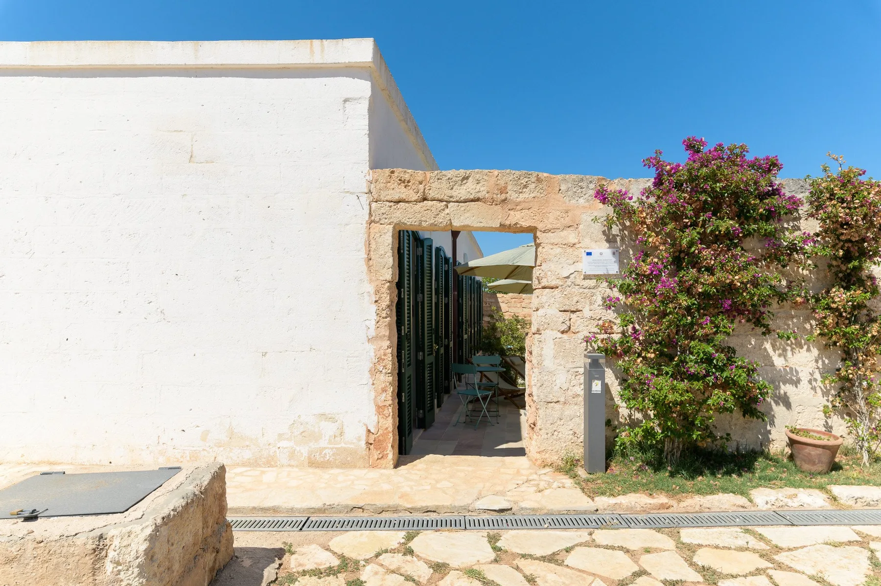 A stone doorway opens onto a shaded patio, with flowering shrubs beside it.