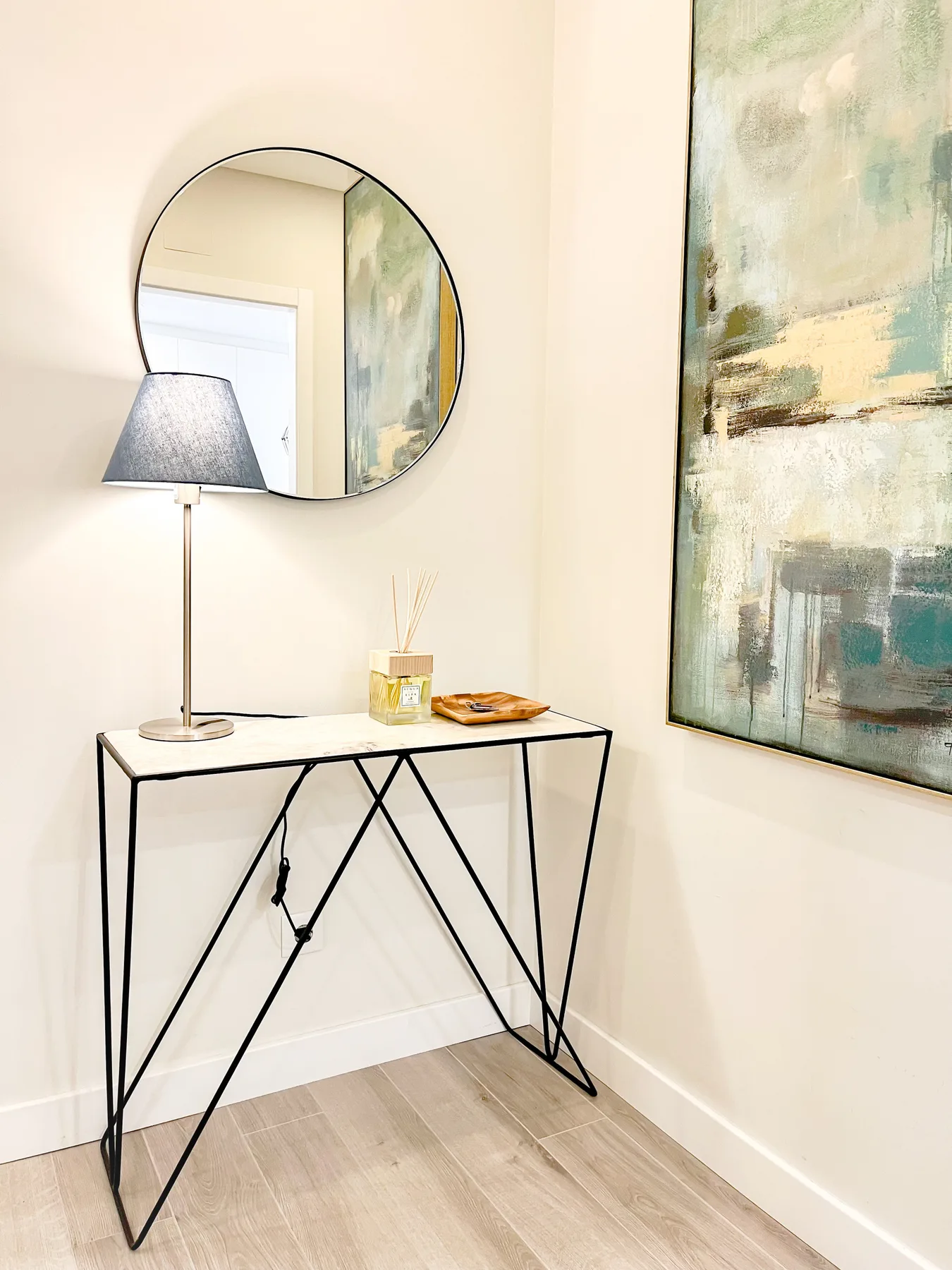 A narrow console table with a lamp and round mirror stands beside an abstract painting.