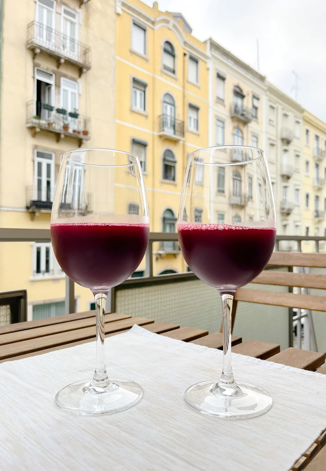 Two glasses of red wine sit on a balcony table overlooking nearby buildings.