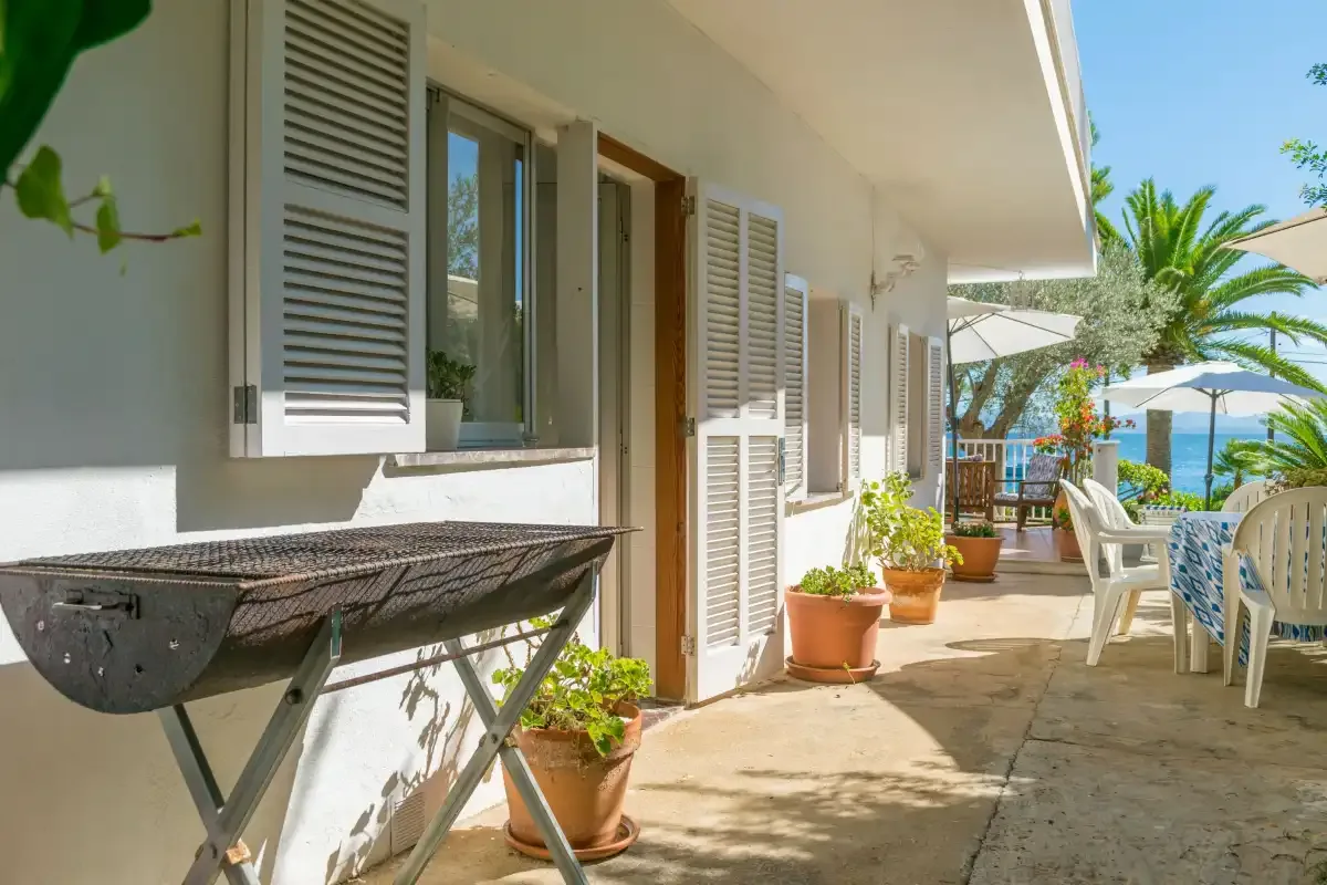 Una terraza exterior soleada junto a un edificio blanco, con plantas y asientos junto a la piscina.