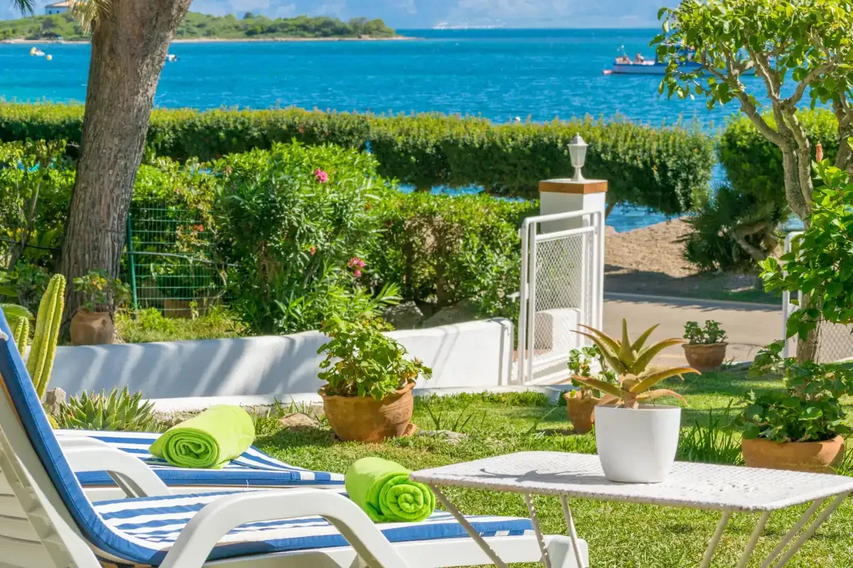 Una zona de jardín con asientos da a una bahía turquesa, con tumbonas y plantas en macetas.