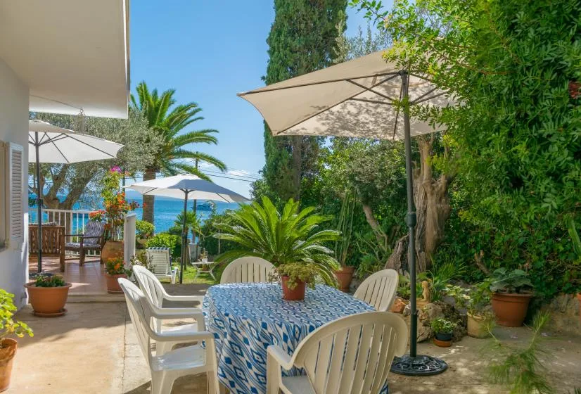 Un comedor al aire libre con mesa, sillas y sombrillas entre plantas de jardín.