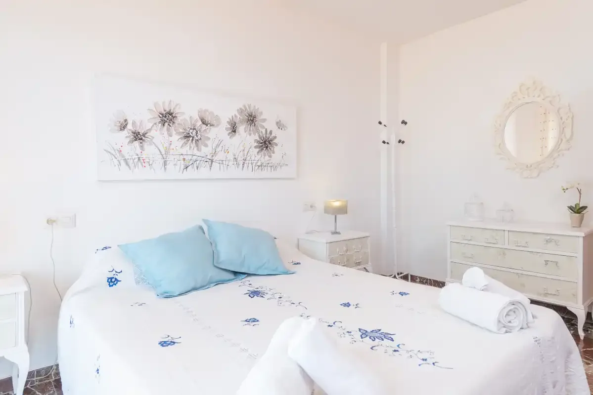 Un dormitorio luminoso con colcha floral, almohadas azul claro y muebles claros.