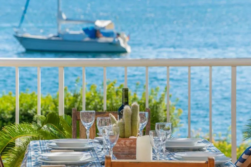 Una mesa de comedor junto al mar, con copas, está junto a una barandilla frente al agua azul.