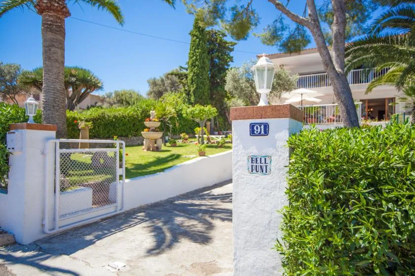 Entrada ajardinada con una puerta blanca, vegetación y un edificio al fondo.