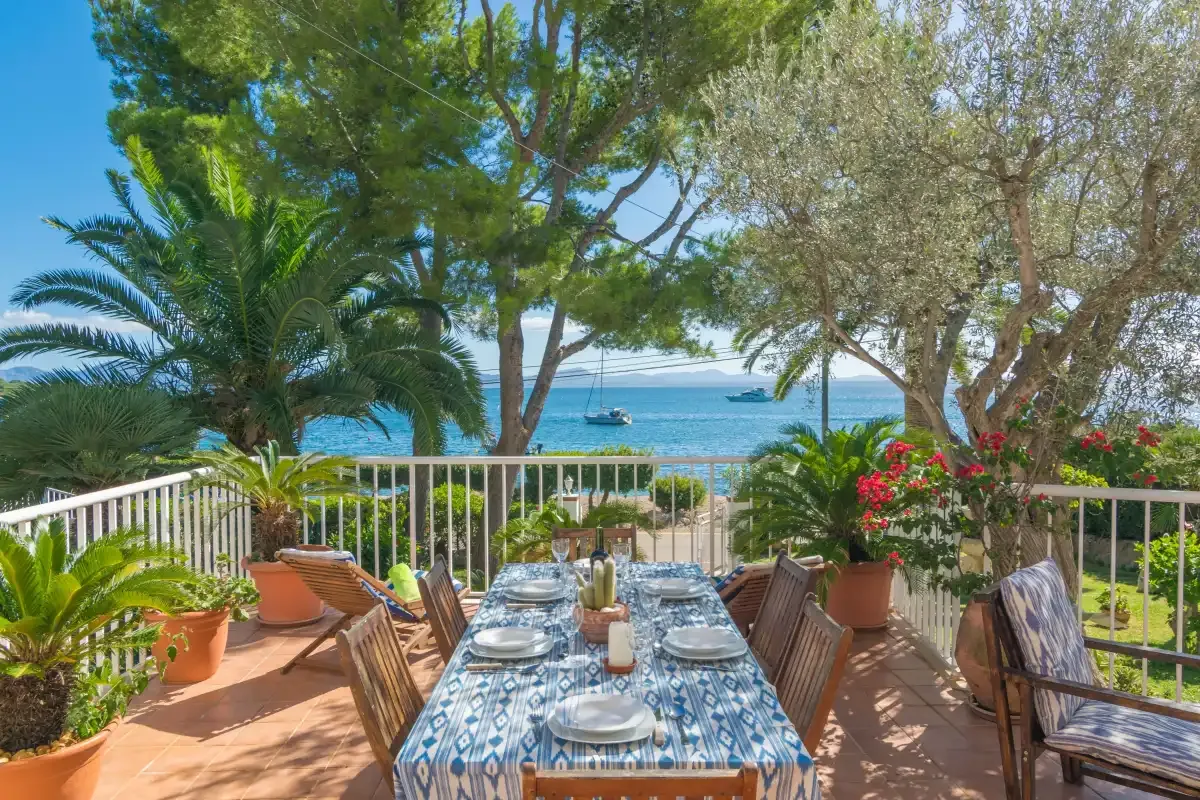Una terraza junto al mar tiene una mesa de comedor al aire libre rodeada de plantas y árboles.