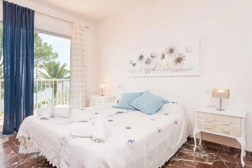 Un dormitorio luminoso con una cama blanca y almohadas azules junto a una puerta de balcón con cortinas.
