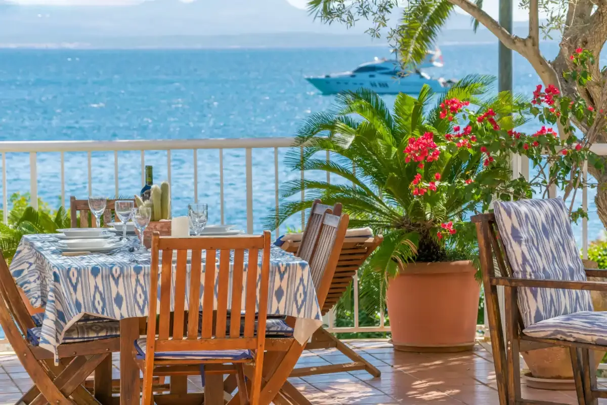 Una terraza junto al mar tiene una mesa de comedor, asientos de madera y plantas en macetas frente al agua.