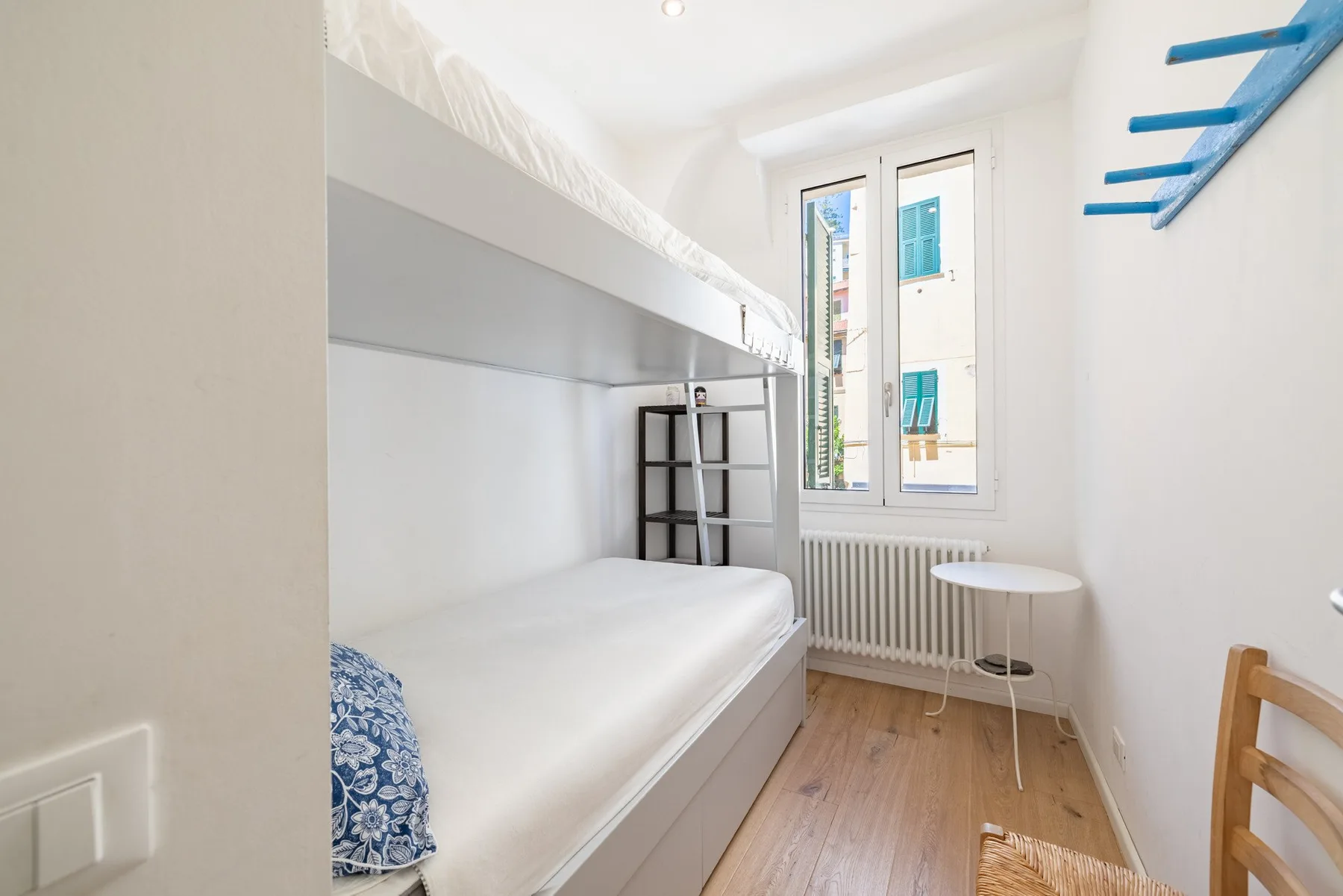 A bright narrow bedroom with a single bed beneath a raised bunk and tall windows.