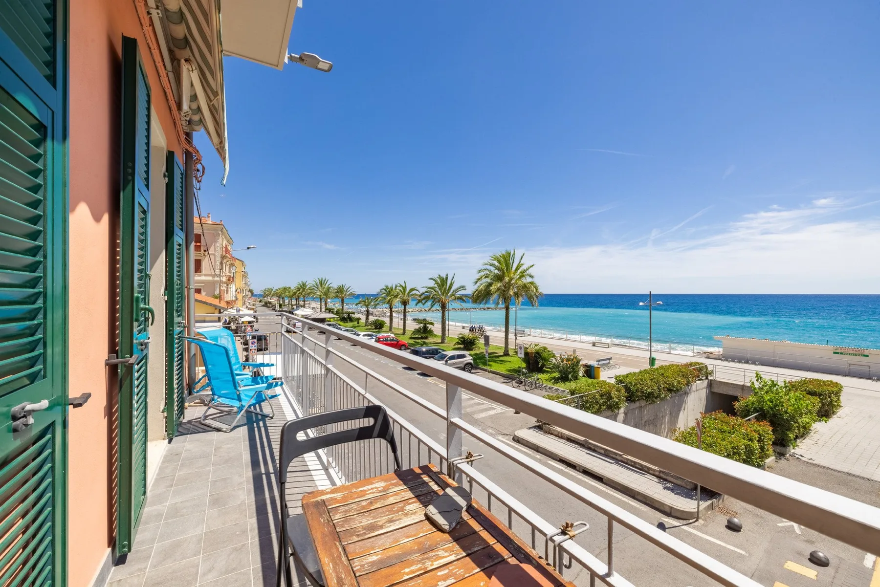 A balcony overlooks a coastal promenade, palm trees, and the sea.
