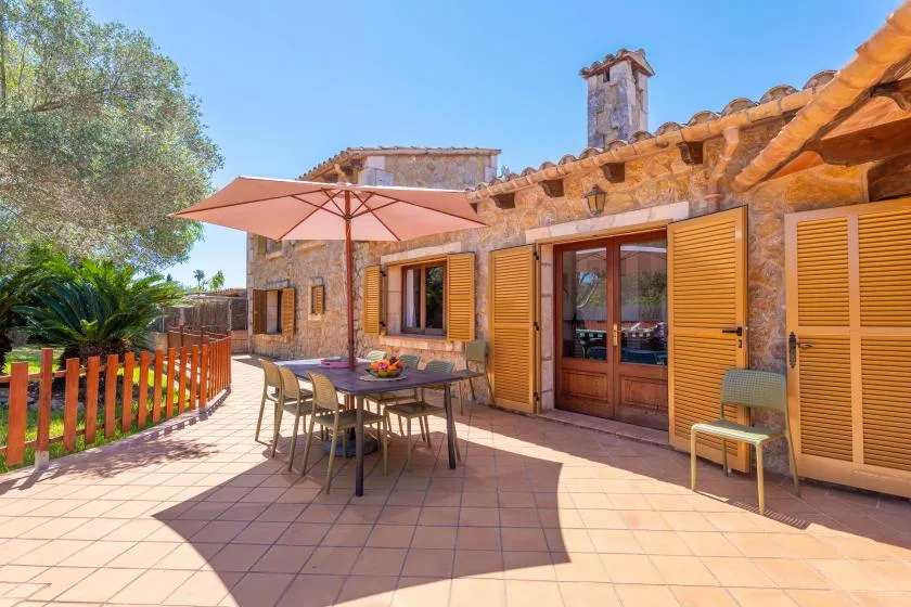 A stone house opens onto a tiled patio with outdoor seating and an umbrella.