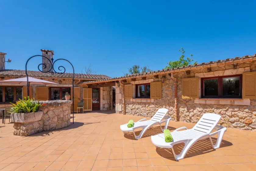 A sunny stone courtyard features two white lounge chairs and potted plants.