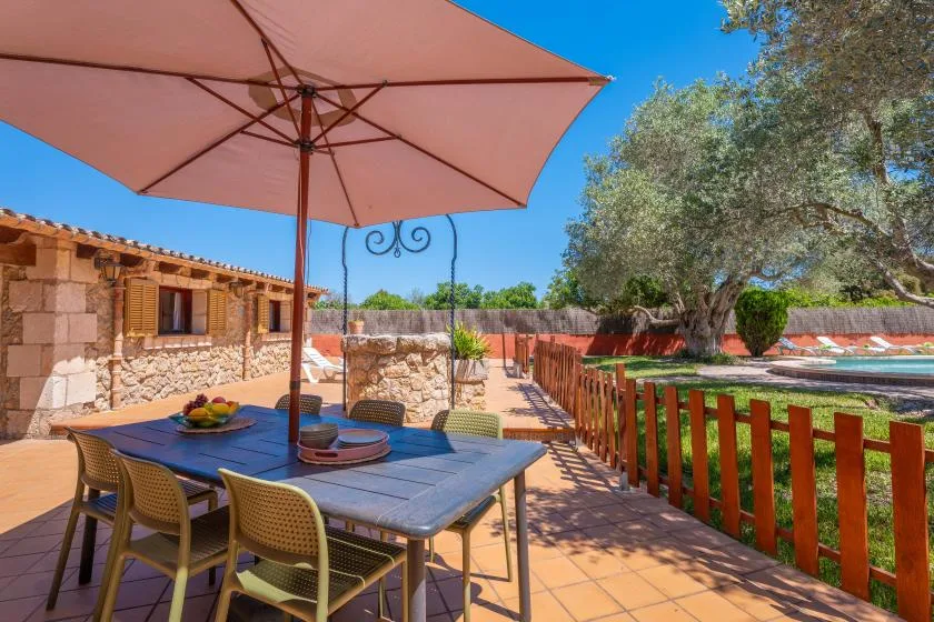 A tiled outdoor patio has a dining table under a large umbrella beside a fenced pool.