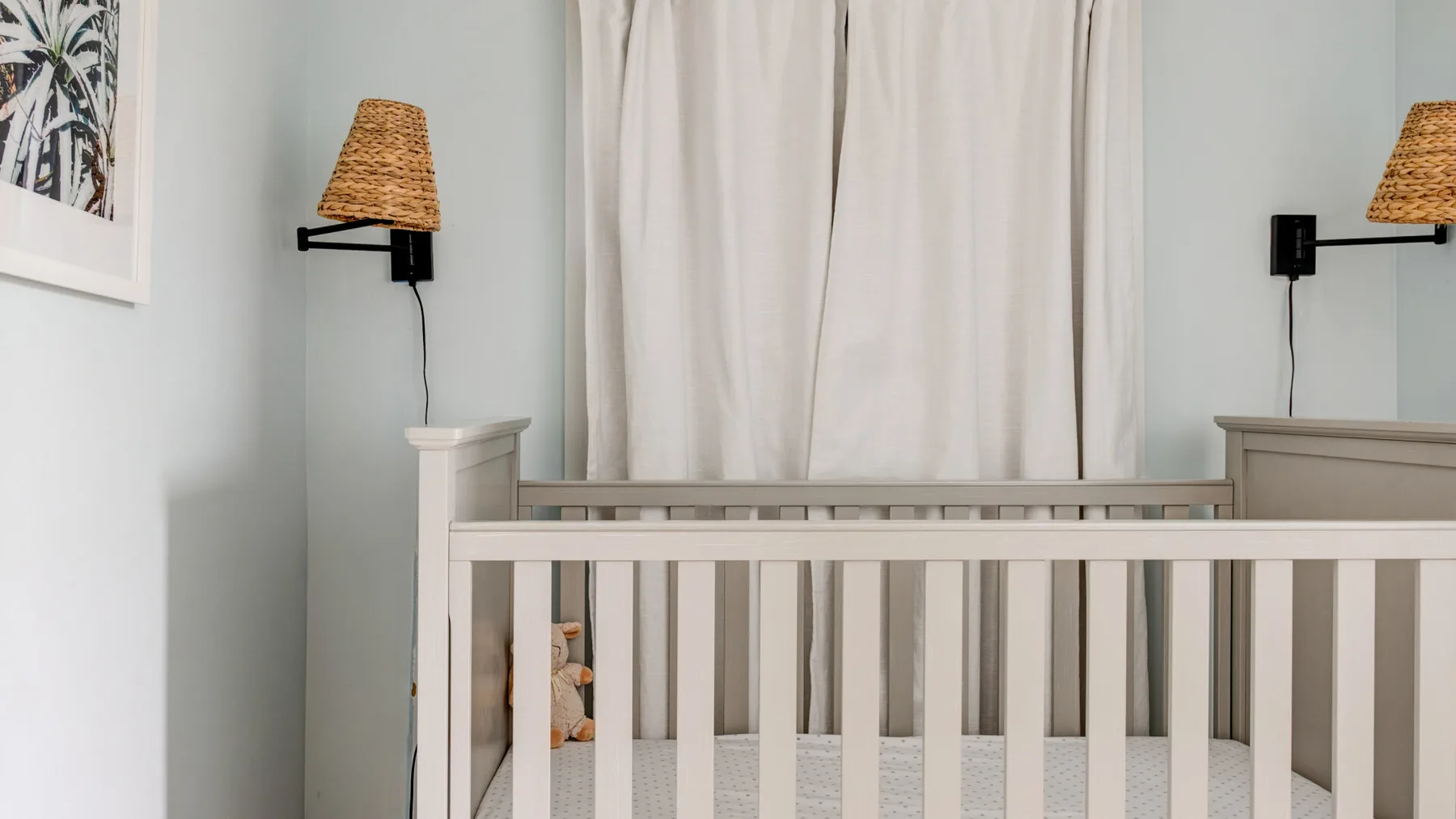 A nursery with a crib in front of light curtains and wall-mounted lamps.