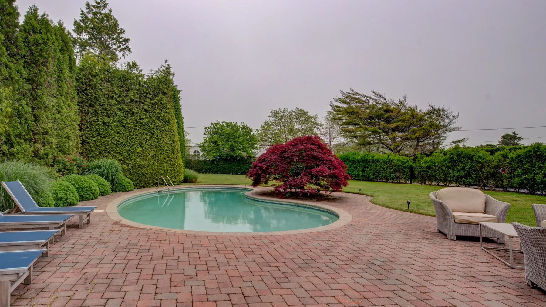 A swimming pool sits in a paved patio surrounded by greenery.