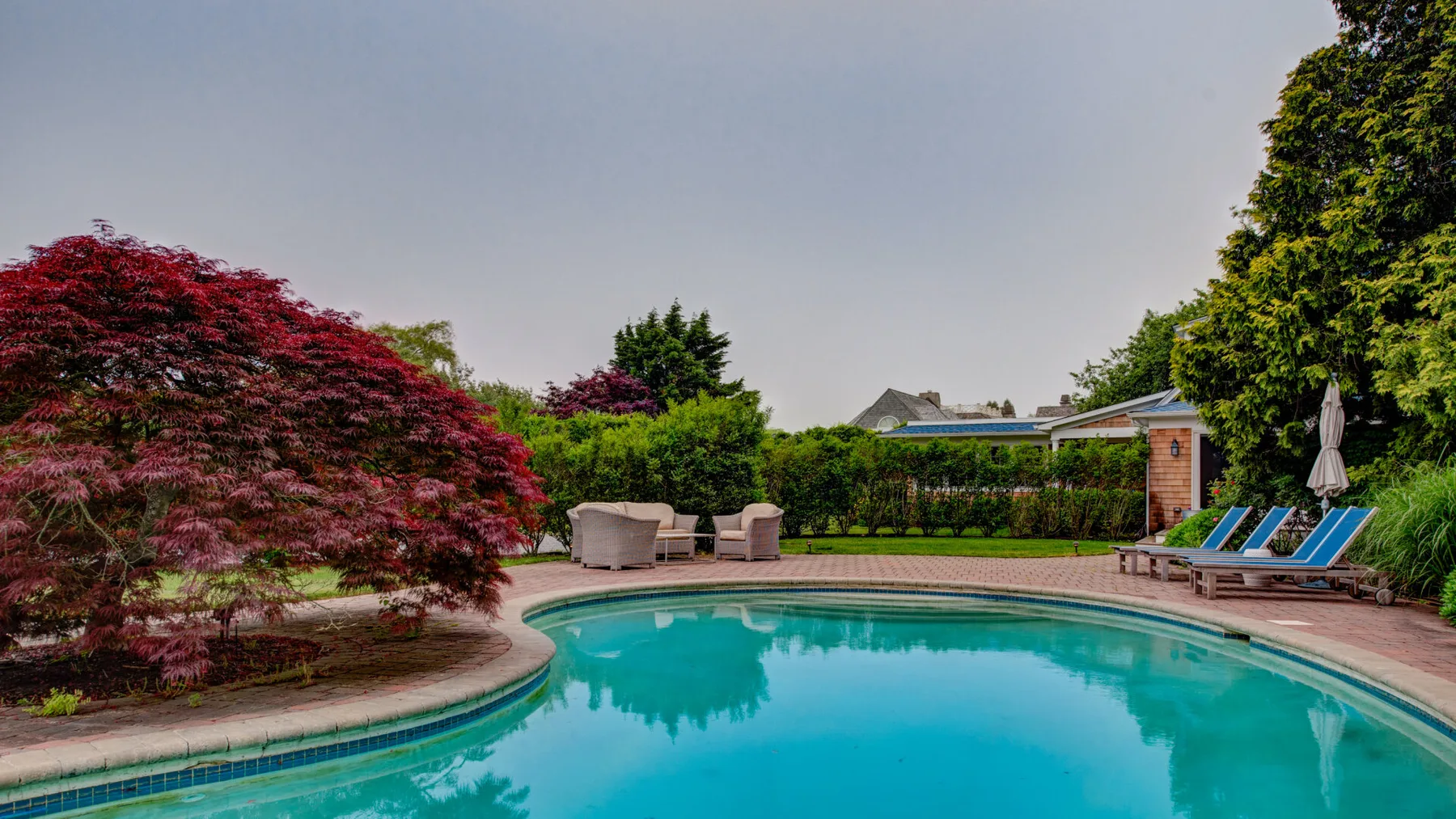 Una piscina al aire libre está rodeada de árboles frondosos y asientos de jardín.