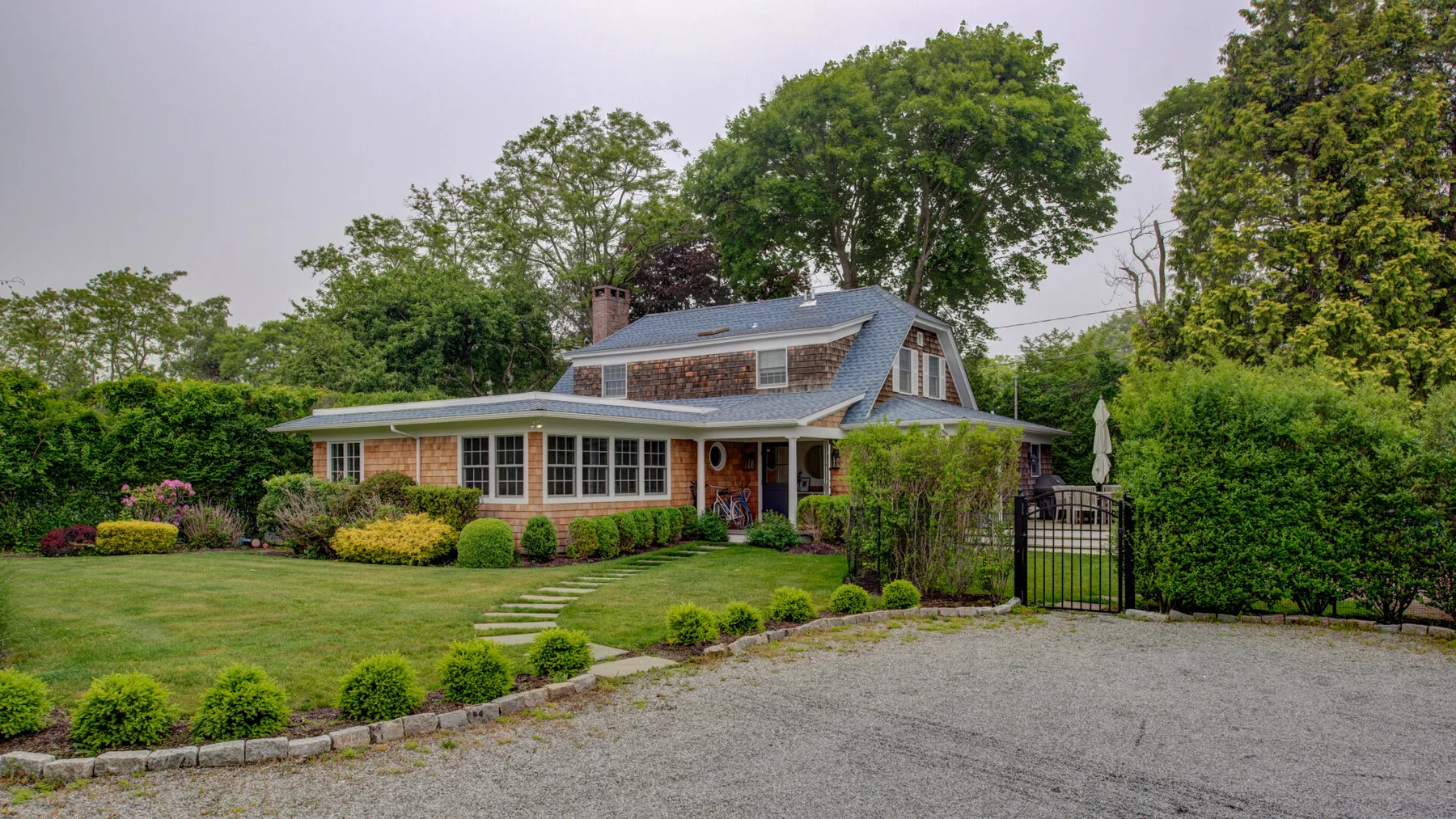 A brick house sits beside a gravel driveway, with trees and landscaped gardens.