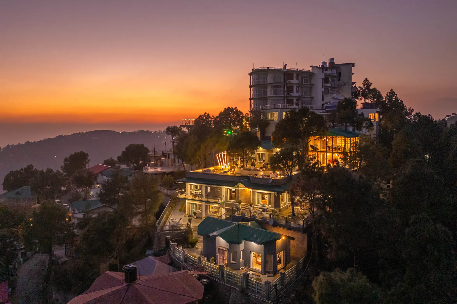 A hillside building complex glows with lights against an orange sunset sky.