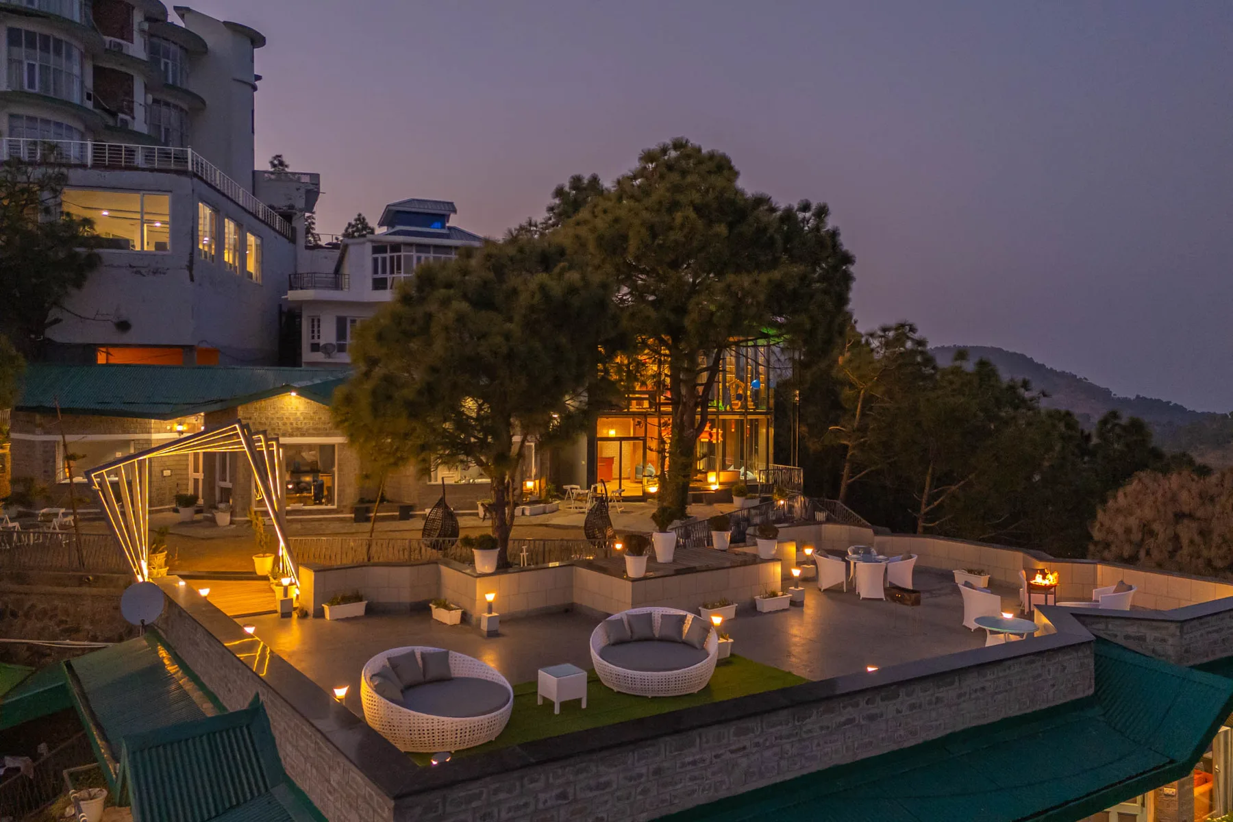 An outdoor terrace at dusk has seating, trees, and warmly lit buildings.