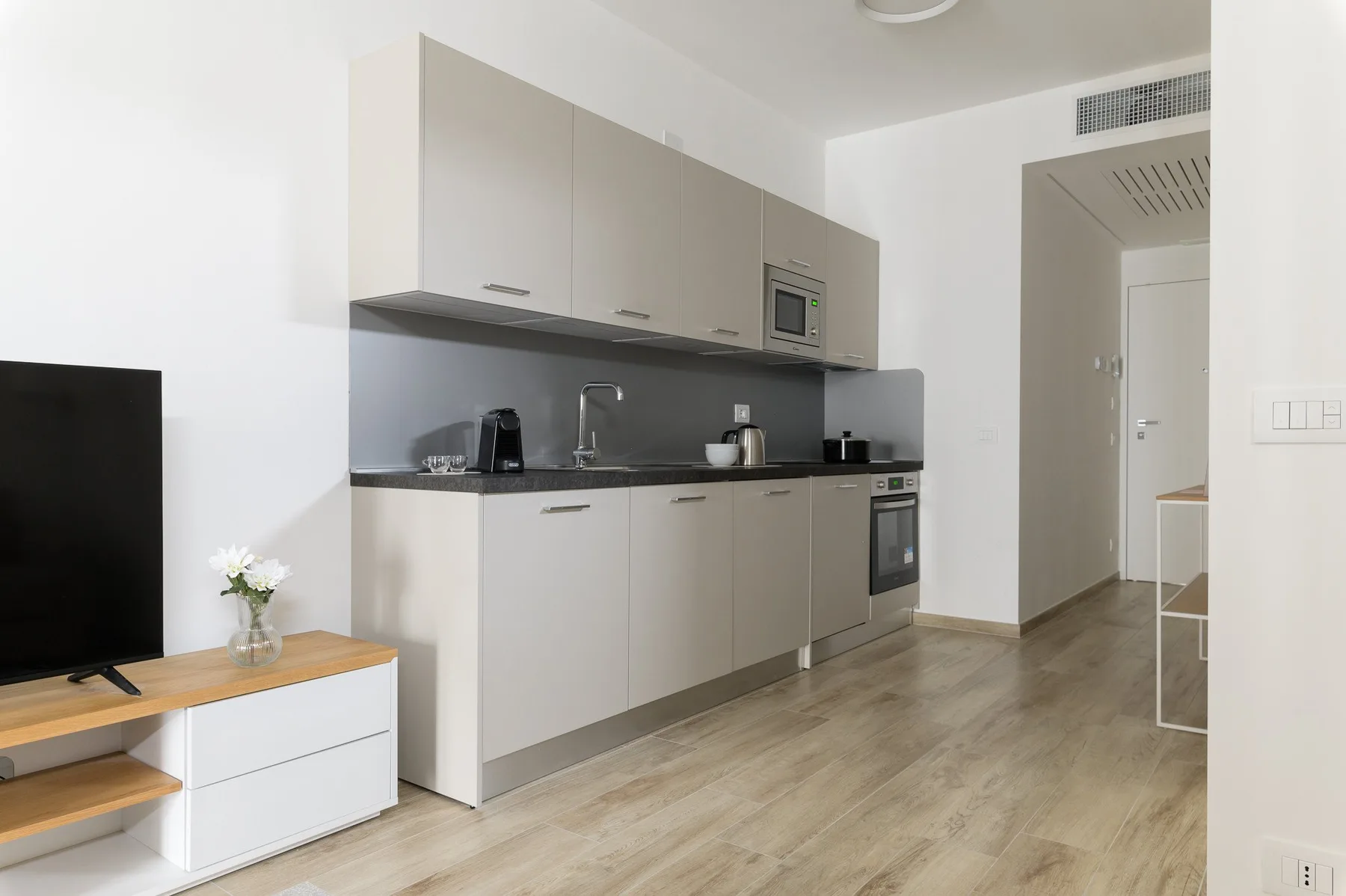 A modern kitchen with light cabinetry, dark countertops, and a television nearby.